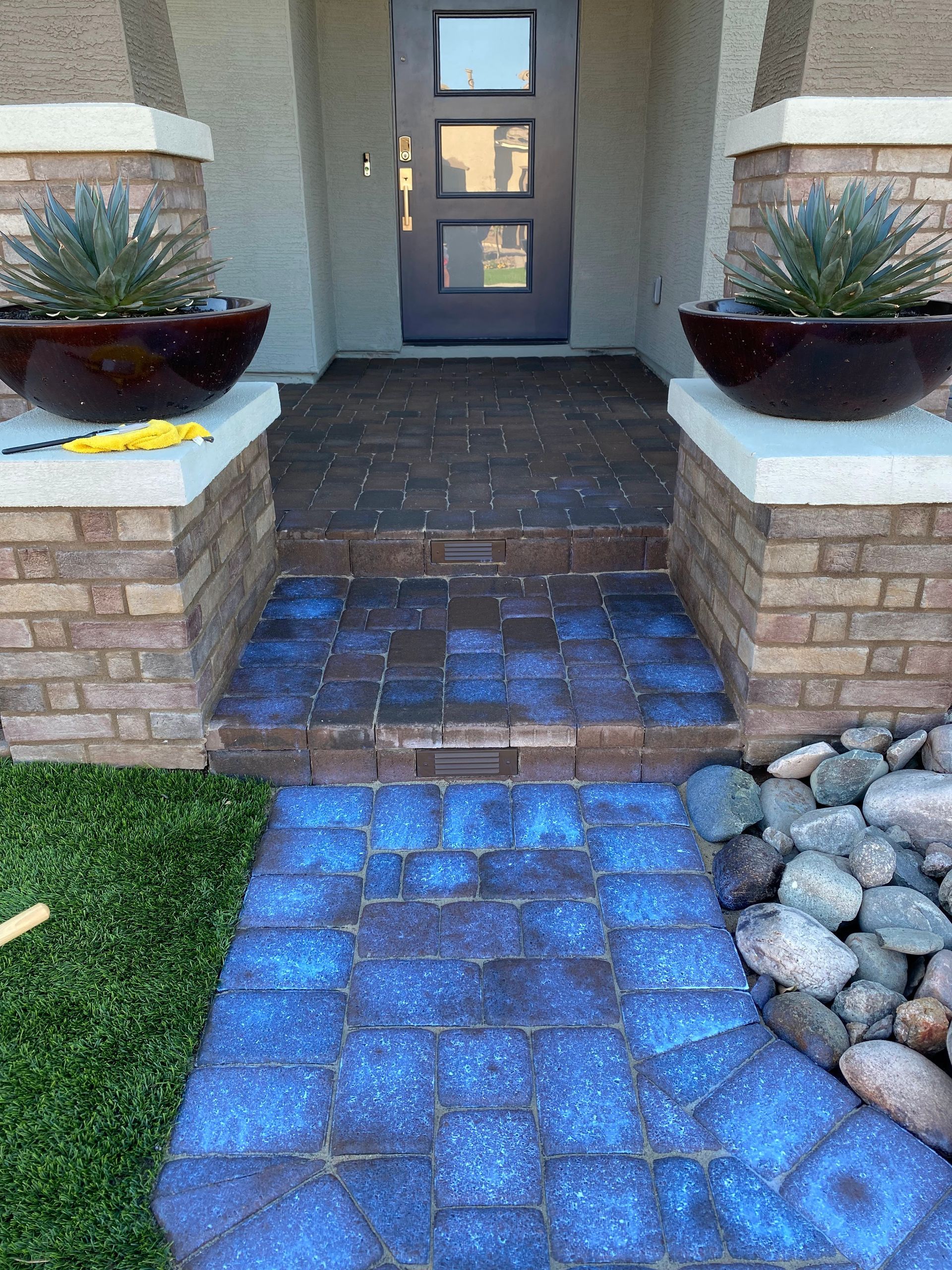 Brick pathway and steps leading to a dark front door, flanked by planters atop brick columns.