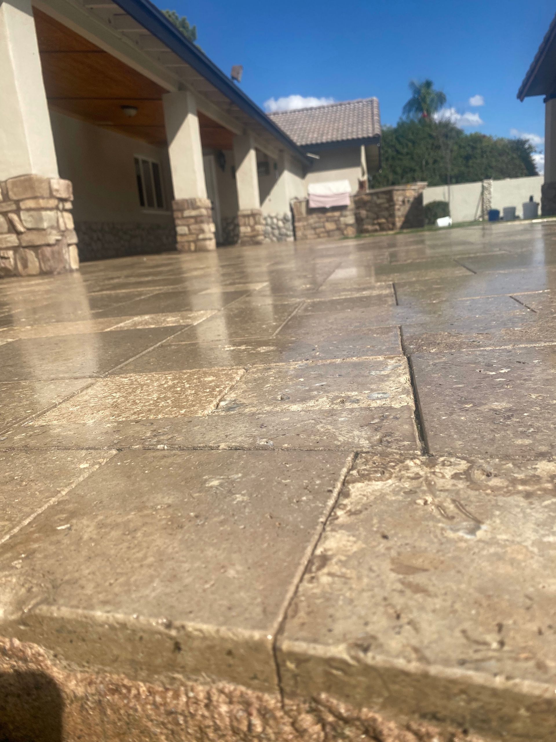 Beige stone patio reflecting sunlight, with a house and blue sky in the background.