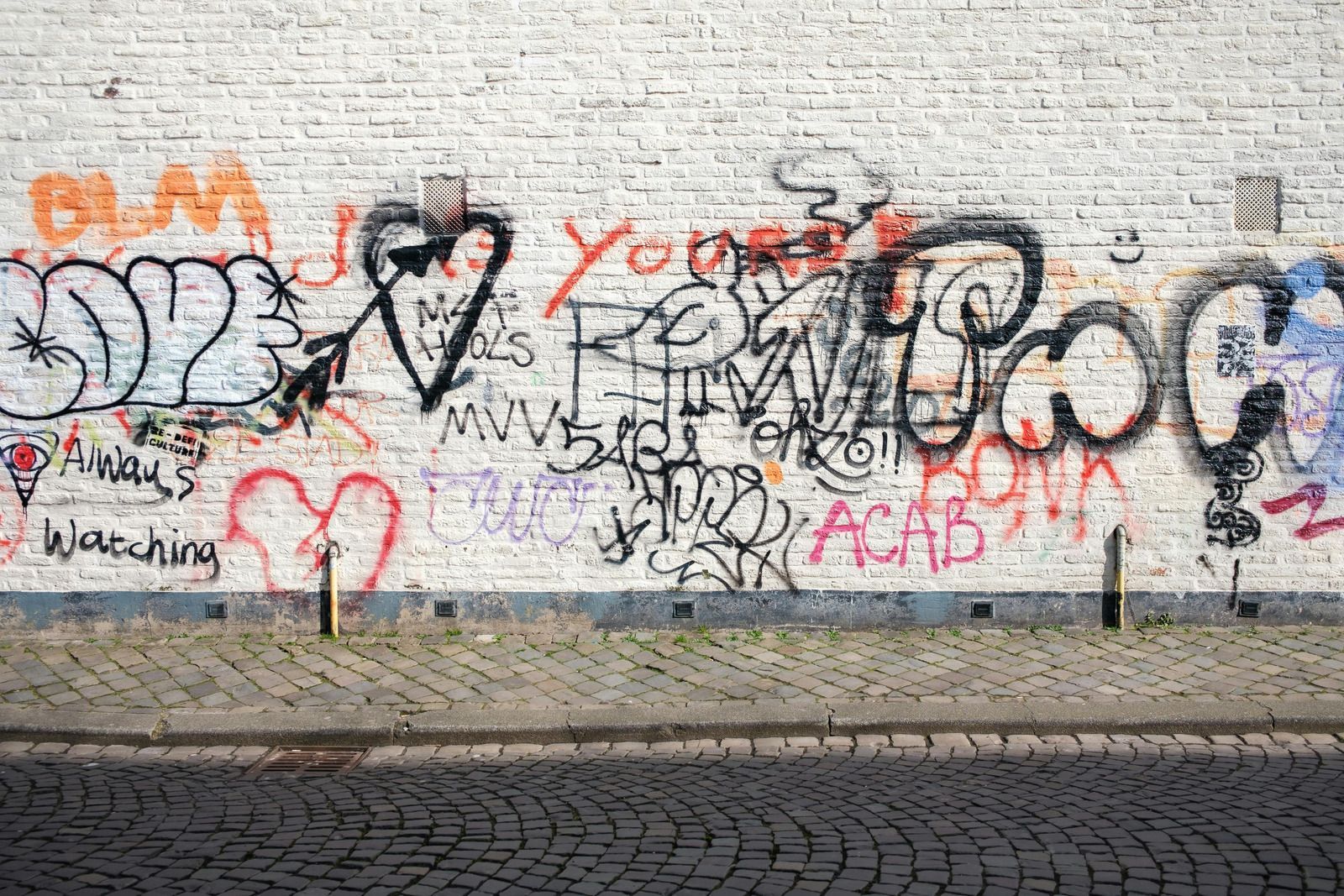 Graffiti on a white brick wall with hearts, words, and symbols; a cobblestone sidewalk in front.