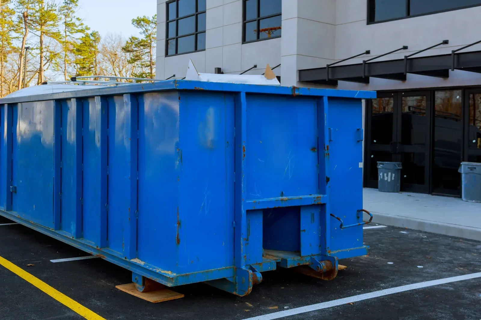 Blue dumpster parked in front of a white building with a black awning and glass doors.