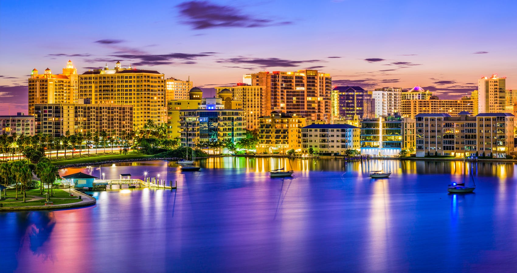 Sunset city skyline with lit waterfront buildings reflecting on calm blue water