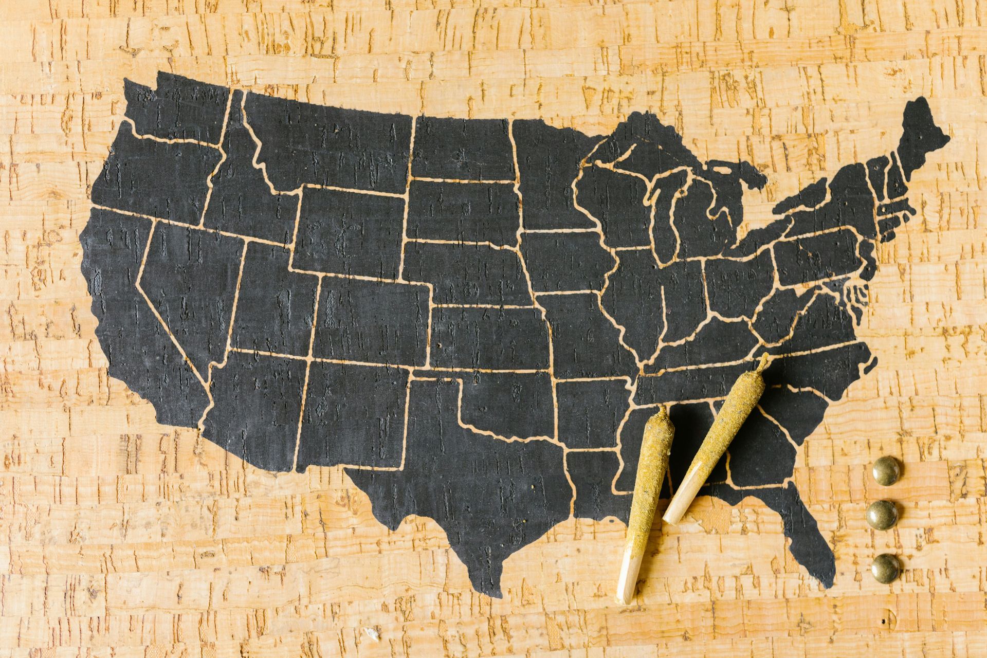 Black outline of the United States map with two cannabis joints and three metal tacks on a wood background.