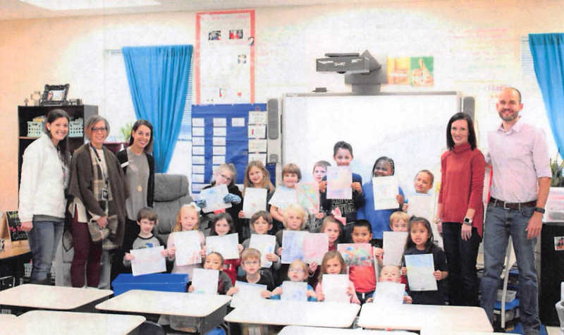 Classroom group photo with children, teachers, and adults standing behind desks, smiling at the camera.