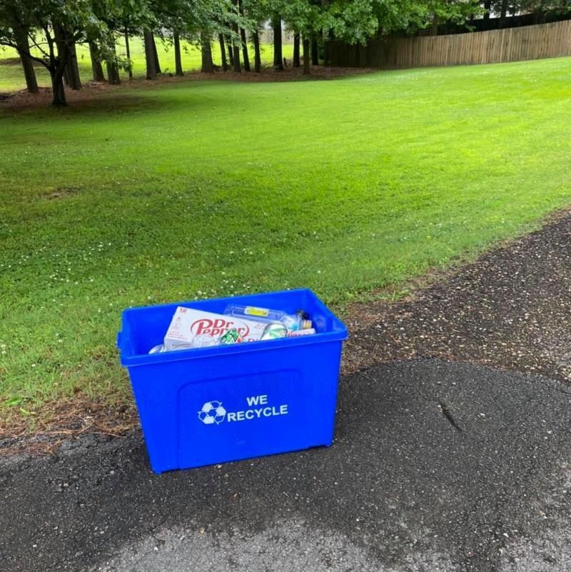 Blue recycling bin on a paved path beside a grassy park lawn and trees.