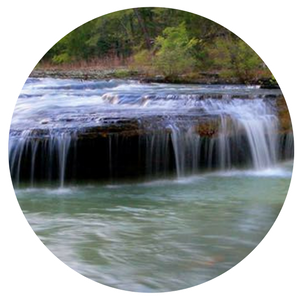 Small waterfall flowing over a rocky ledge into a calm green pool, surrounded by trees