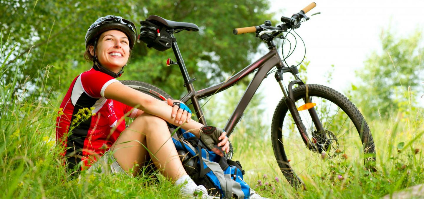 Cyclist in red helmet and shirt sits on grass beside mountain bike in a sunny field