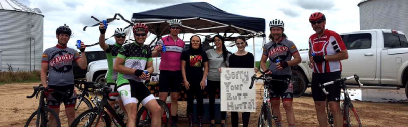 Cyclists posing beside bikes under a canopy at an outdoor event, with a support truck in the background.