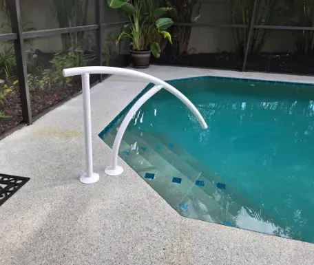 White handrail next to swimming pool steps. Pool with turquoise water, gray patio.