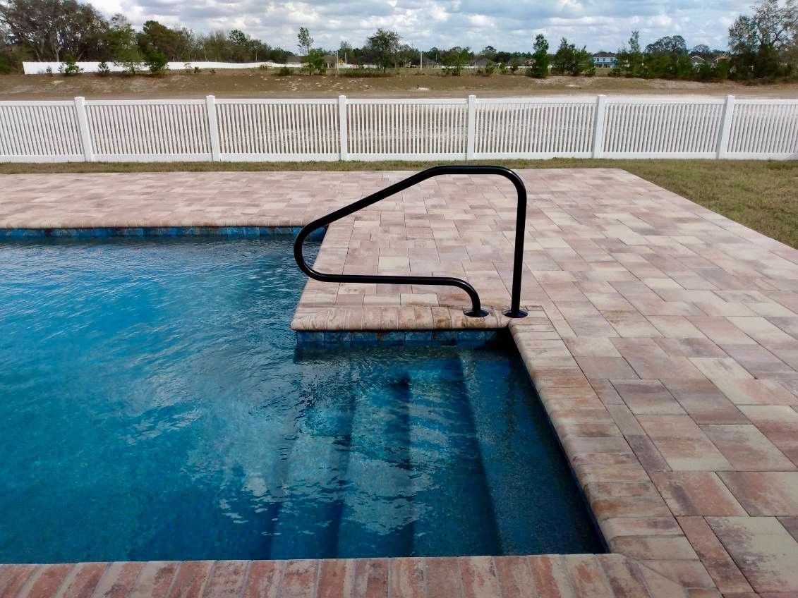 Pool with brick patio and a handrail for the stairs. White fence and trees are in the background.