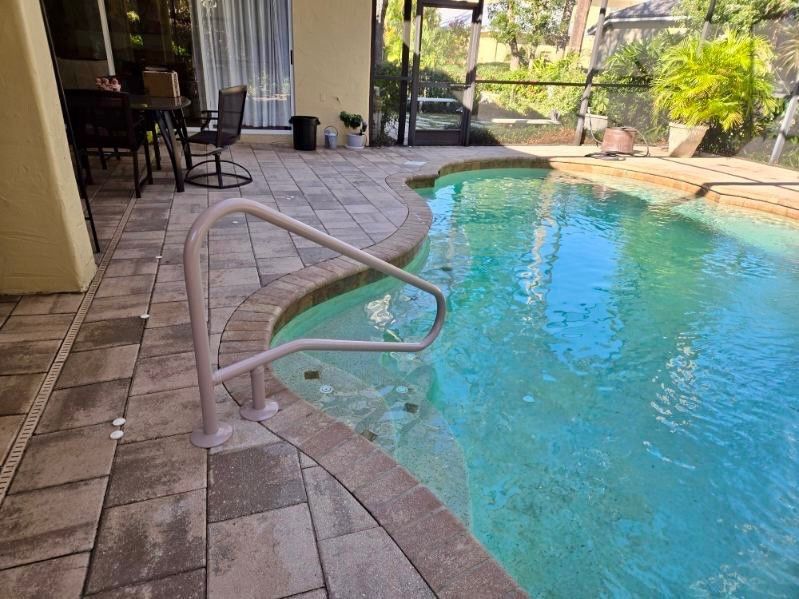 Poolside with brick paving and a curved pool with a handrail; sunny outdoor scene.