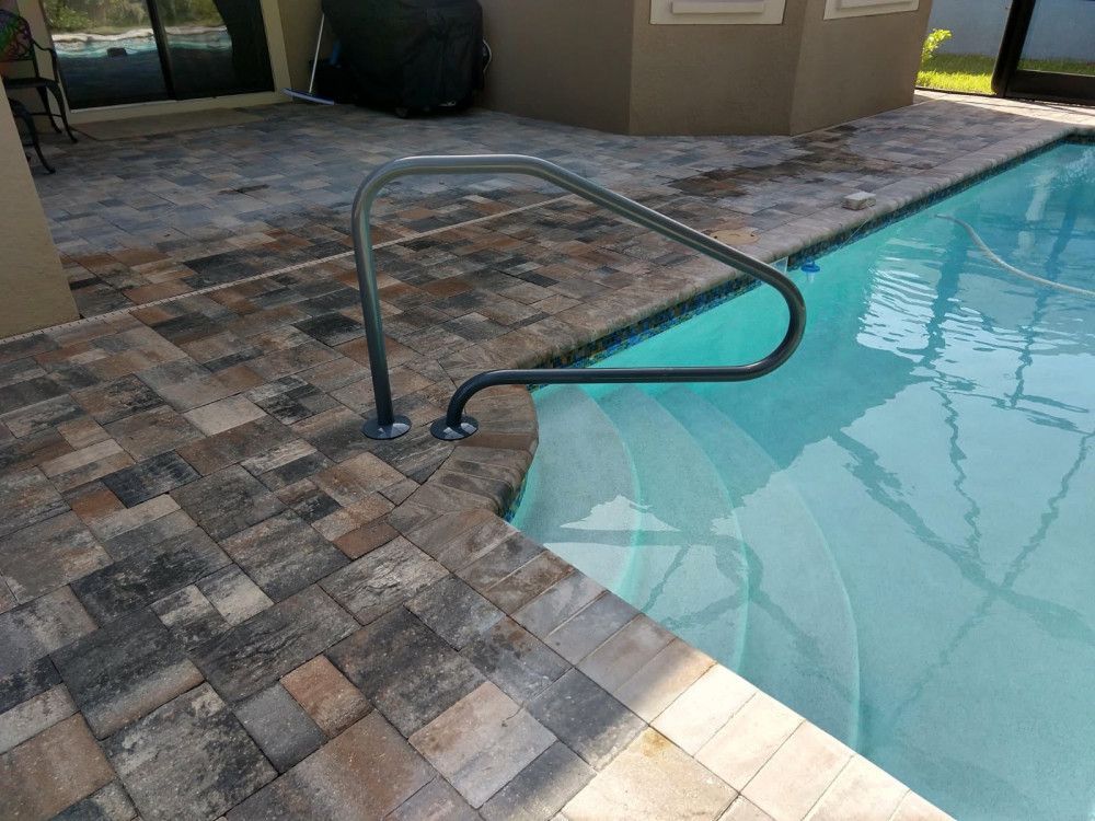 Pool handrail over pool steps. Handrail is dark gray. Paved patio surrounds the pool.