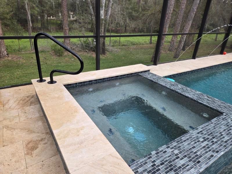 Hot tub and pool with tiled surround, railing, and view of trees beyond a screened enclosure.