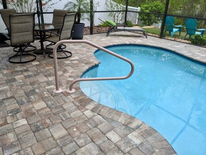 Poolside with a handrail. Brown pavers, blue water. Patio chairs and lounger in the background.