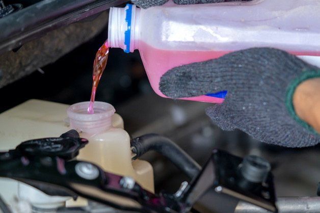 Gloved hand pours pink liquid from a bottle into a car coolant reservoir