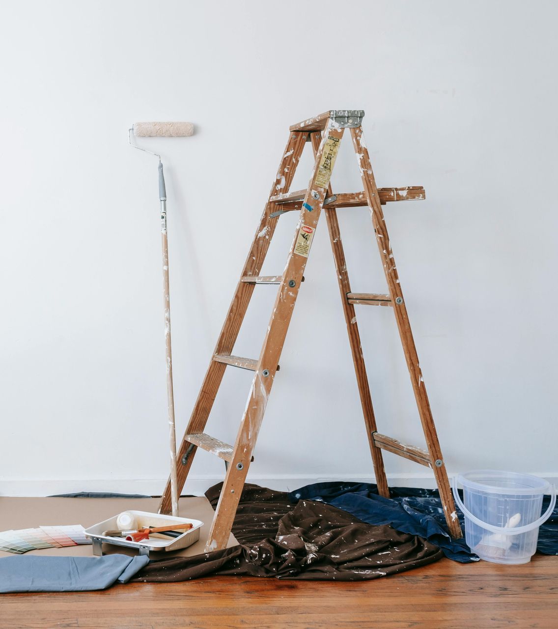 A wooden ladder is sitting on the floor next to a paint roller and bucket.