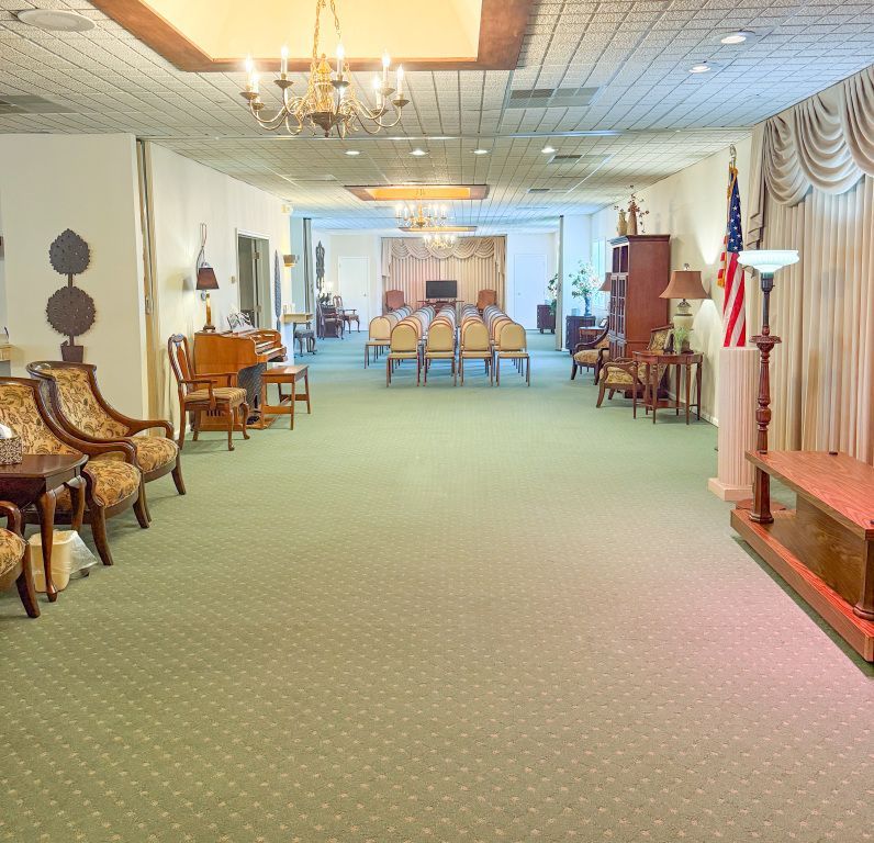 Funeral home interior: green carpet, chairs, casket, chandeliers, American flag, and drapes.
