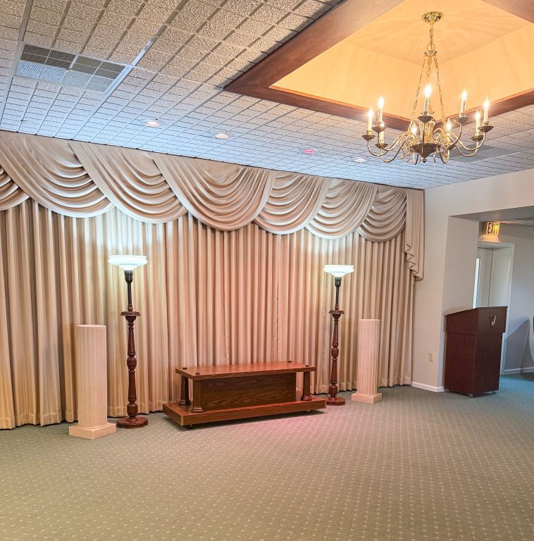 Interior of a beige funeral home with a draped stage, podium, and chandelier.