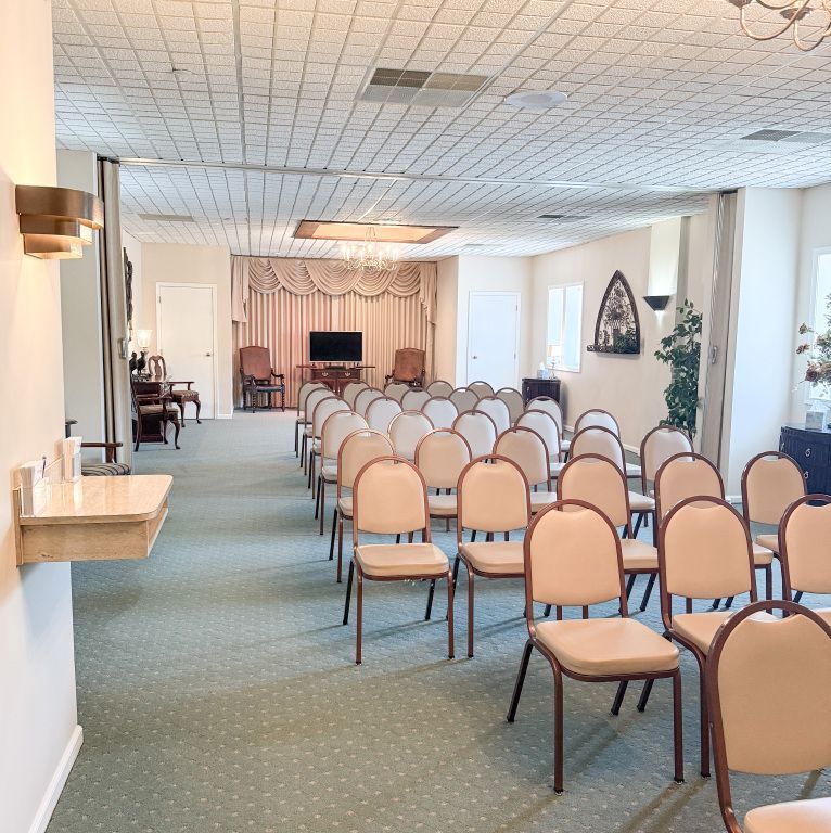 Rows of beige chairs face a small stage in a funeral home. Green carpet, white walls, and a decorative ceiling.