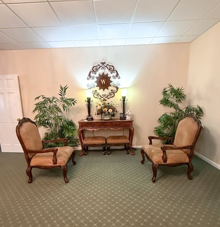 Two ornate chairs flank a console table with a decorative sunburst, lamps, and plants in a room with a green carpet.