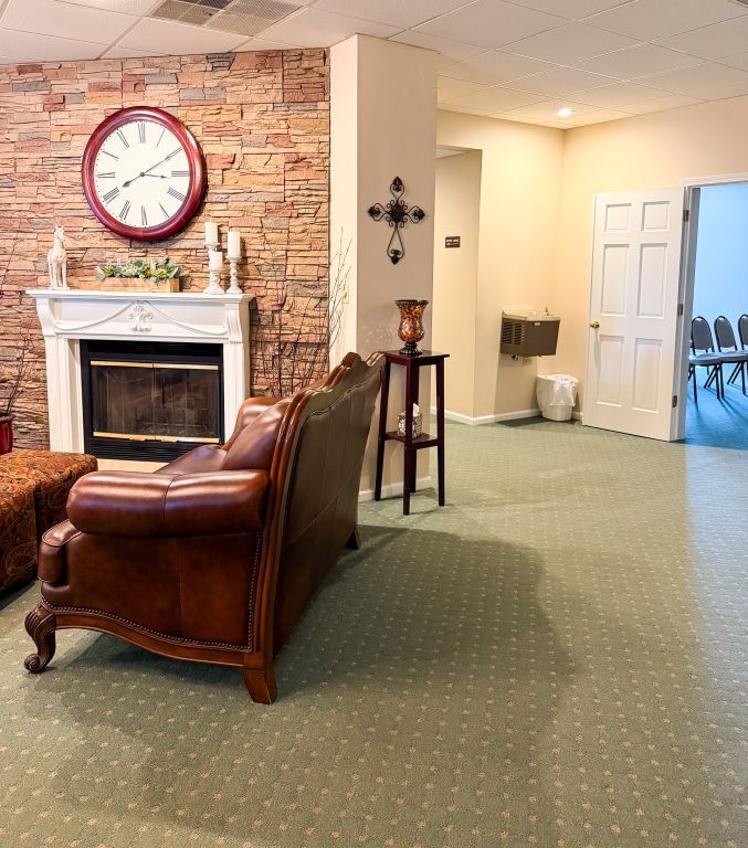 A waiting room with a fireplace, leather couch, clock, and doorway to a seating area.