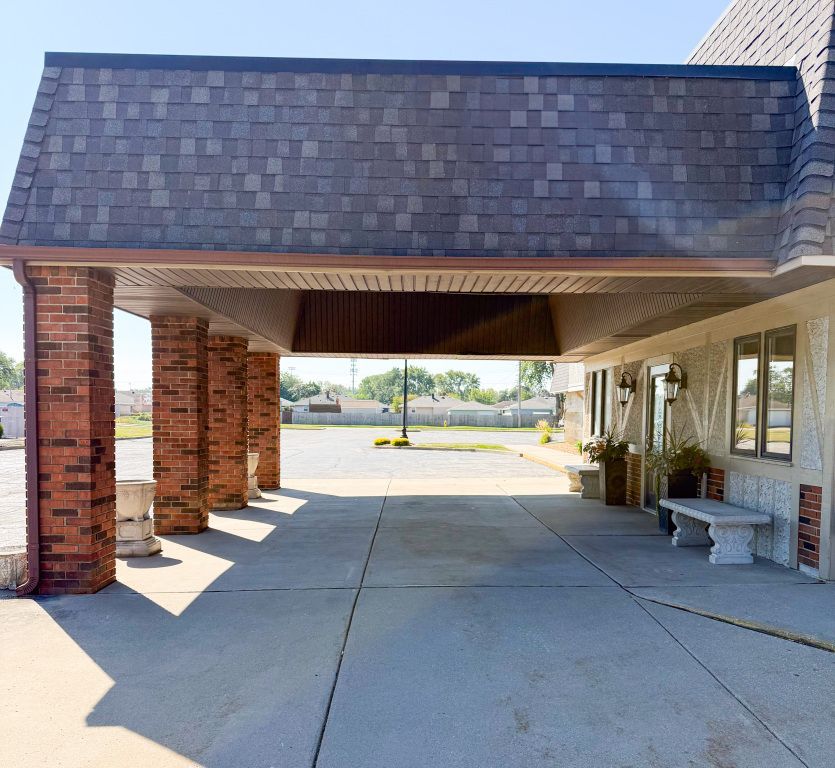 Covered driveway with brick columns. Gray roof. Concrete. Sunny day.