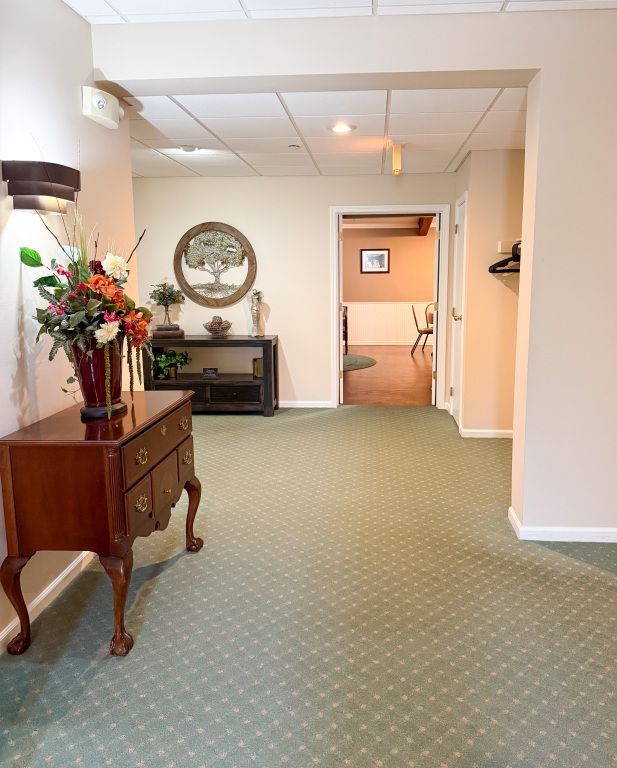 Hallway with green carpet, beige walls, and wood furniture with floral arrangements. Open doorway leads to another room.