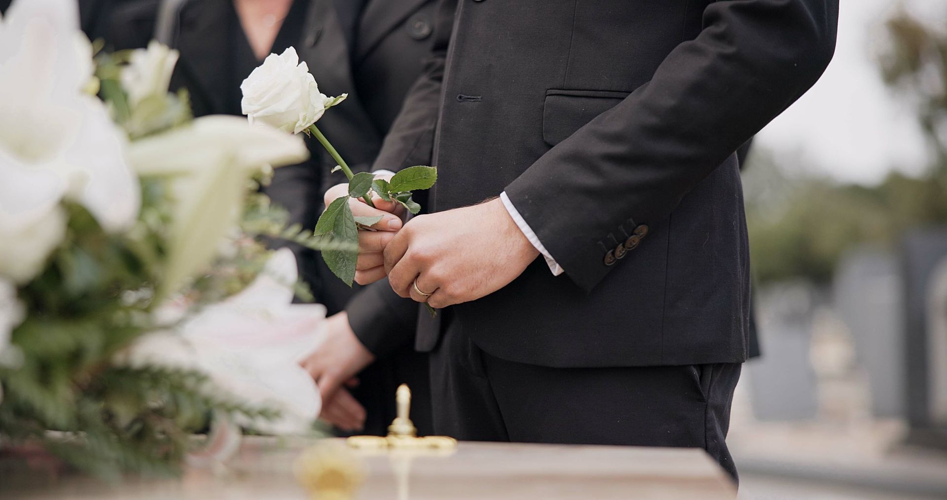 Man in black suit holding a white rose at a funeral, flowers and casket in view.