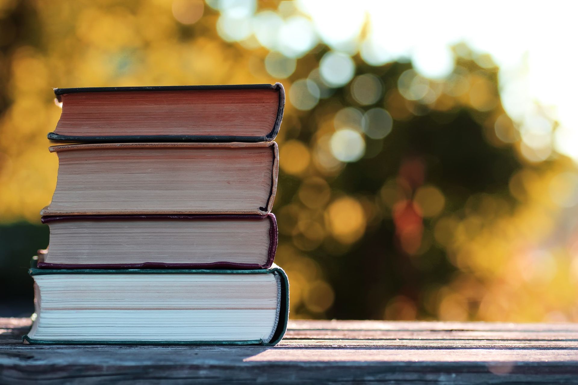 Stack of old books on a wooden surface, sunlight in the background.