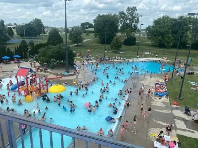Large outdoor public swimming pool crowded with people on a sunny day; slides and green space visible.