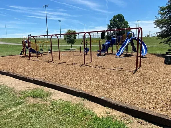 Playground with swings and slides, covered in wood chips, on a sunny day.
