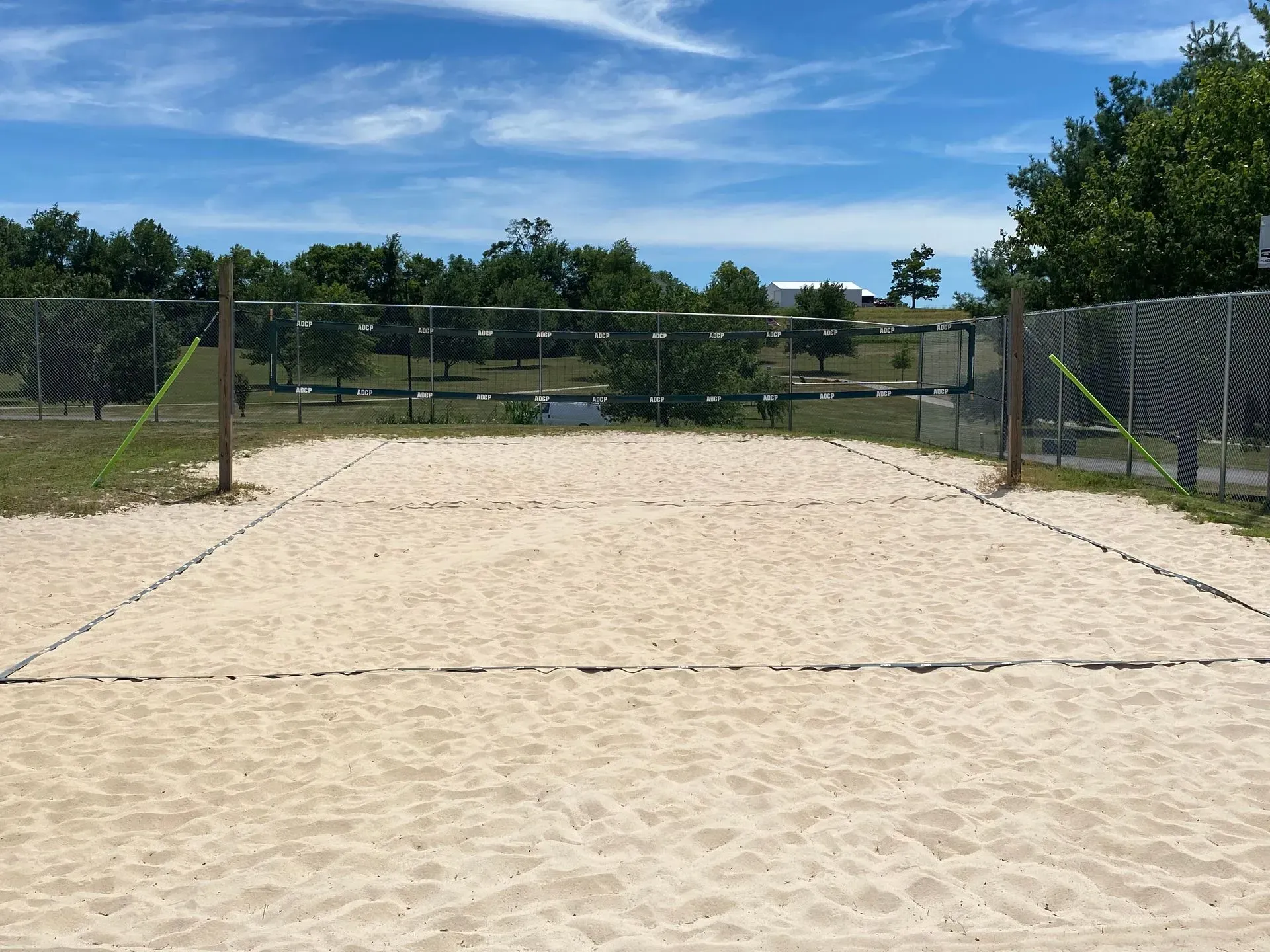A sand volleyball court with a net, empty under a blue sky, surrounded by trees and a fence.