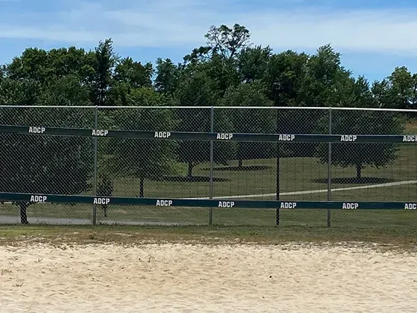 A chain-link fence with green bands, trees and a sandy ground under a blue sky.