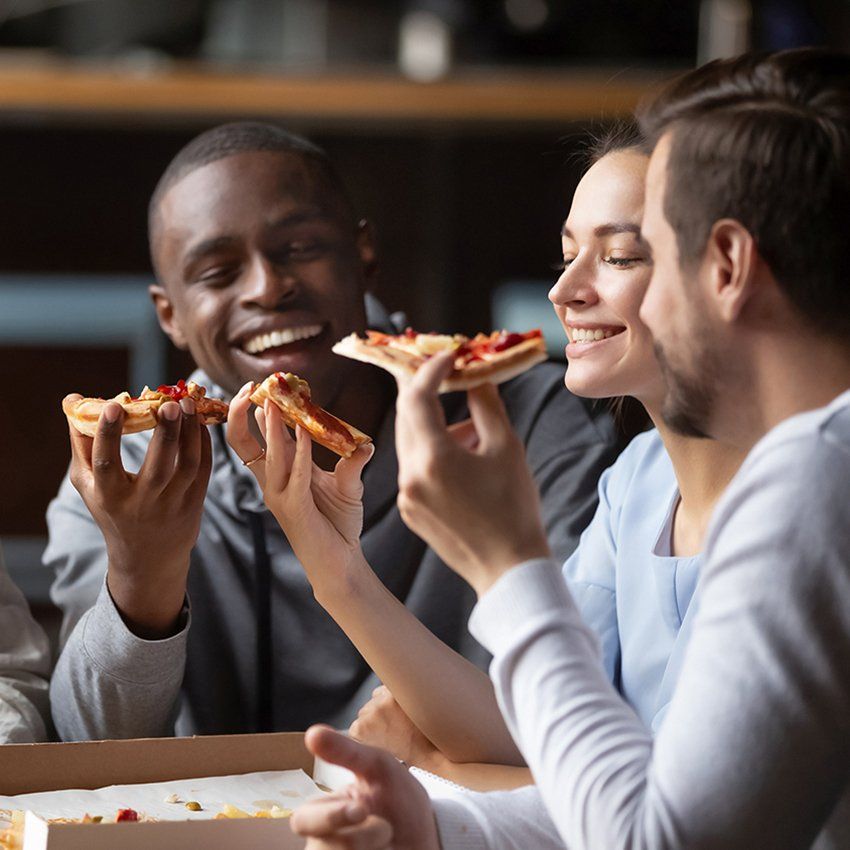Friends smiling and eating pizza slices from a box, indoors.