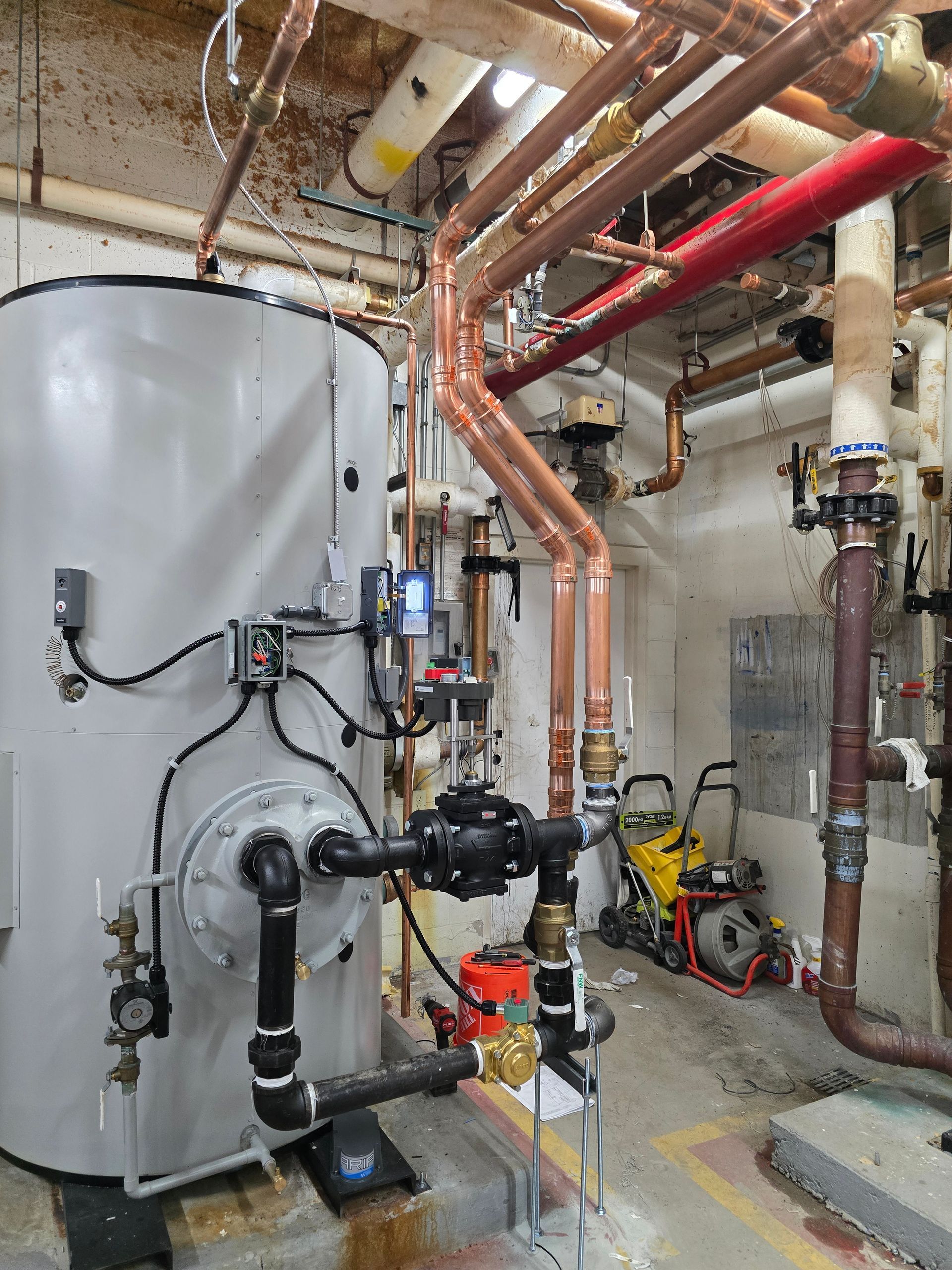 Industrial boiler room with a large white tank, copper pipes, gauges, and maintenance equipment
