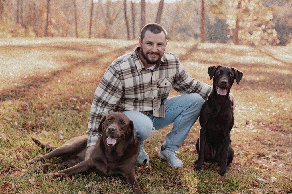 Man kneels outdoors on a leaf-covered path with two dogs, one sitting and one lying down.