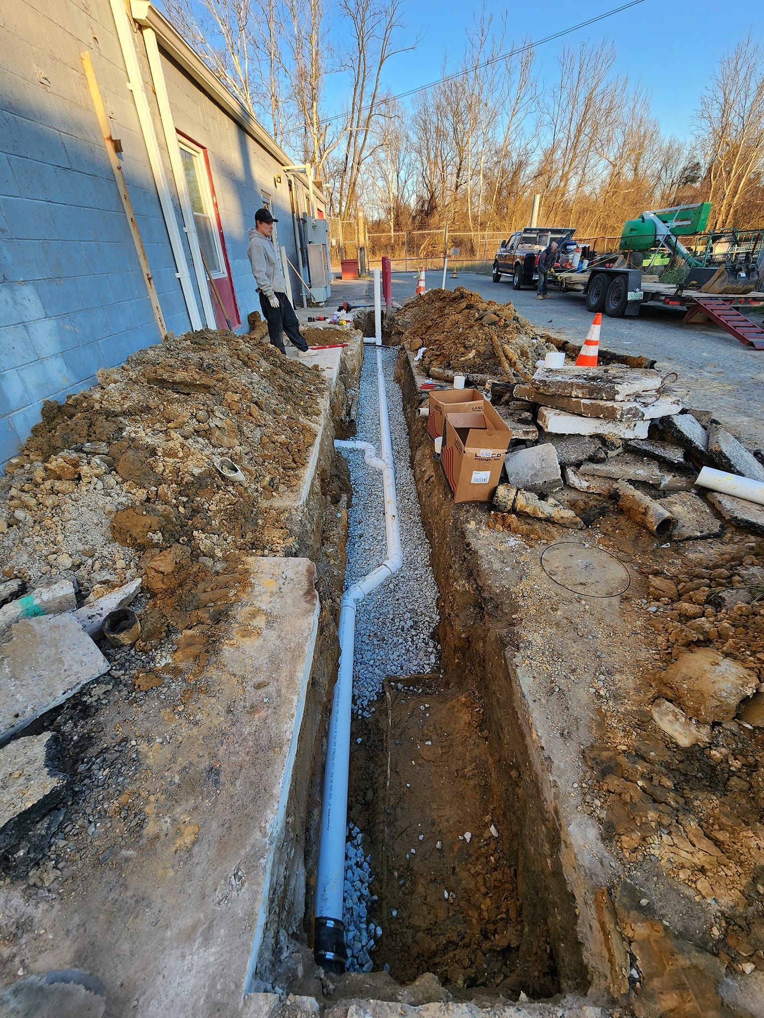 Trench excavation beside a blue building, with workers and muddy construction debris along the sidewalk.
