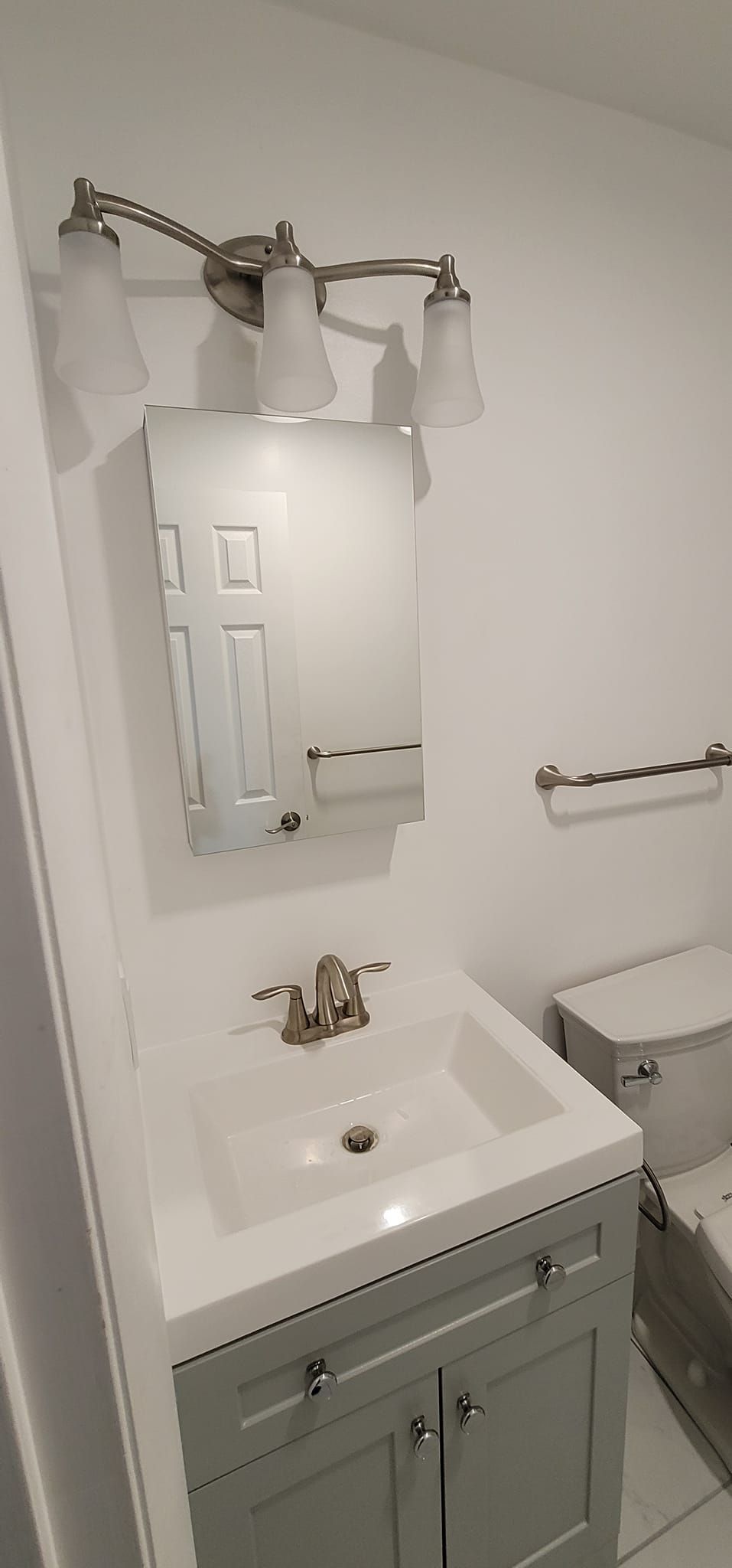Small white bathroom with vanity sink, mirror, light fixtures, and a towel bar beside a toilet.