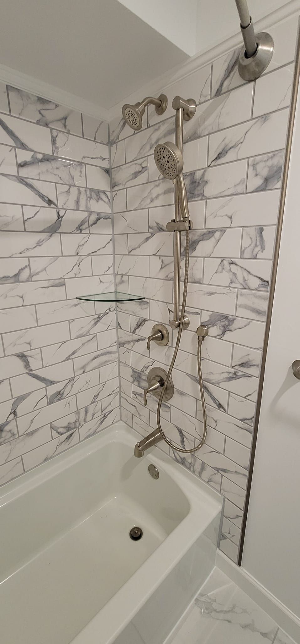 Bathroom shower corner with white tile, black grout, and a tub edge below a brass shower pipe.