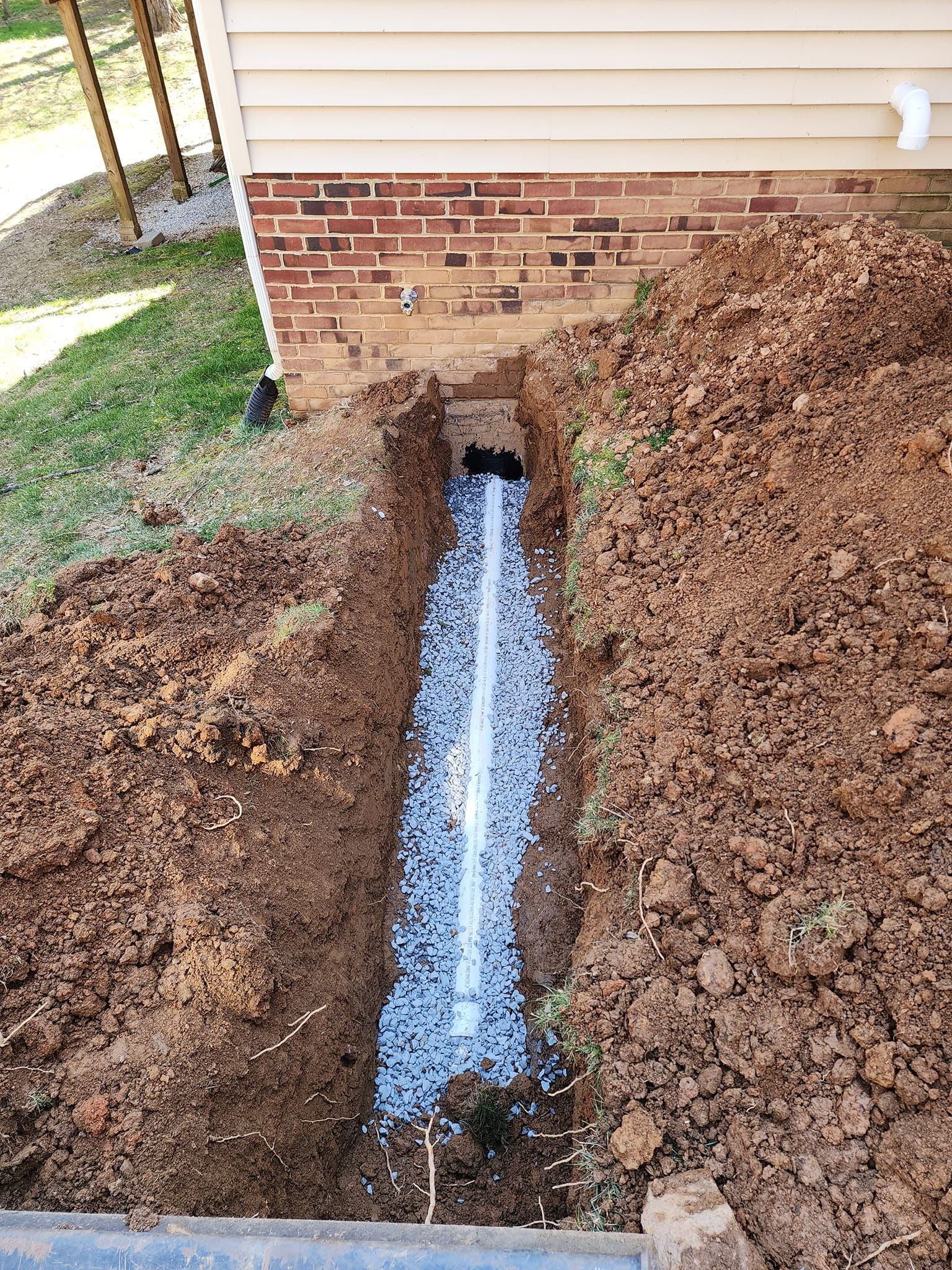 Trench with white insulation foam beside a brick house foundation, surrounded by dirt and leaves.