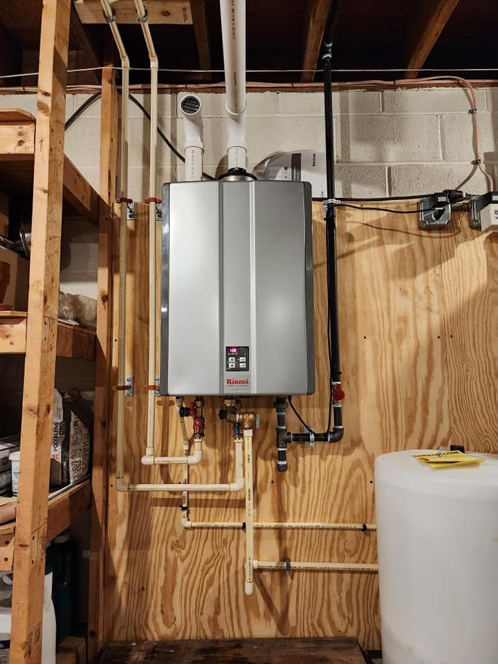 Wall-mounted silver water heater with exposed pipes in a framed utility room, beside a white tank.