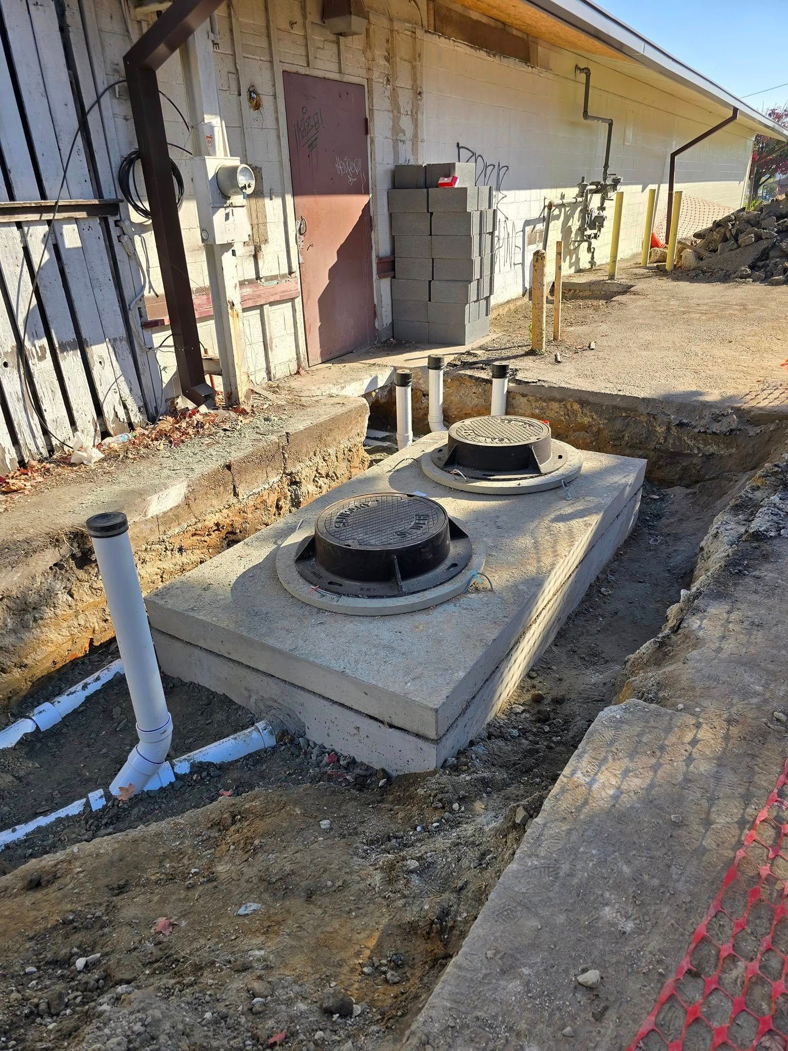 Two utility manholes on a concrete pad beside a building, with construction fencing and gravel around them