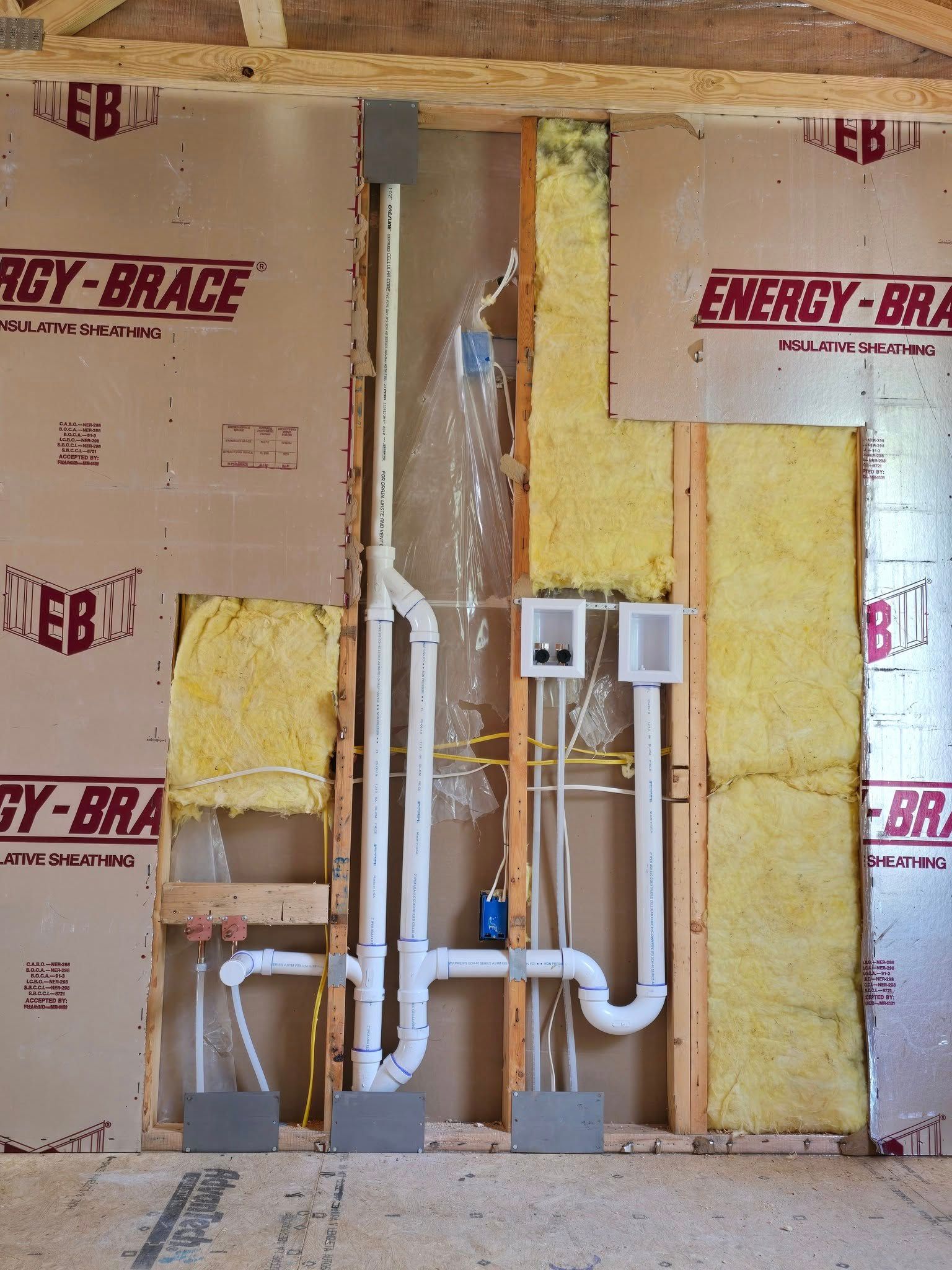 Interior wall under construction with exposed insulation, pipes, wiring, and electrical boxes.