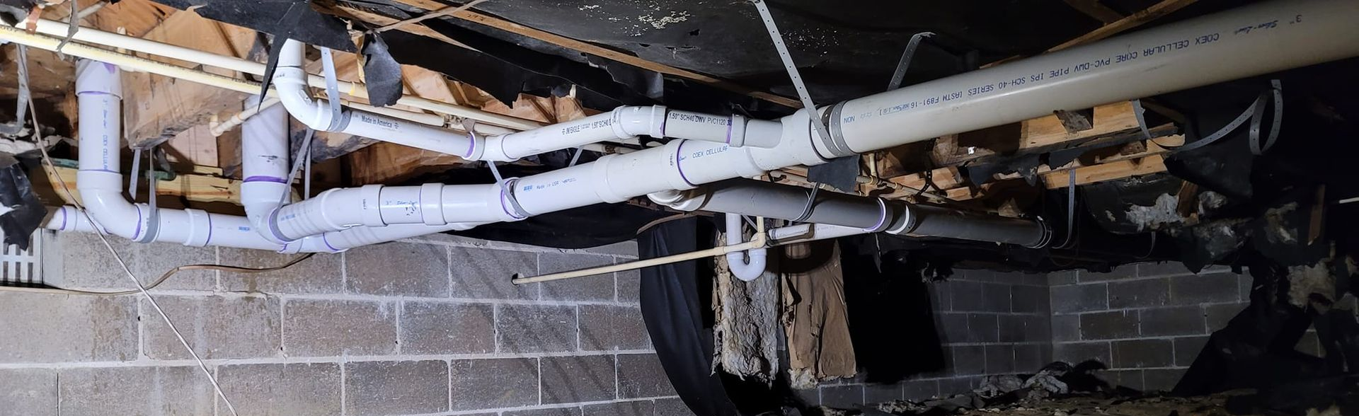 Damaged basement ceiling with exposed pipes, wiring, and debris over a cinderblock wall