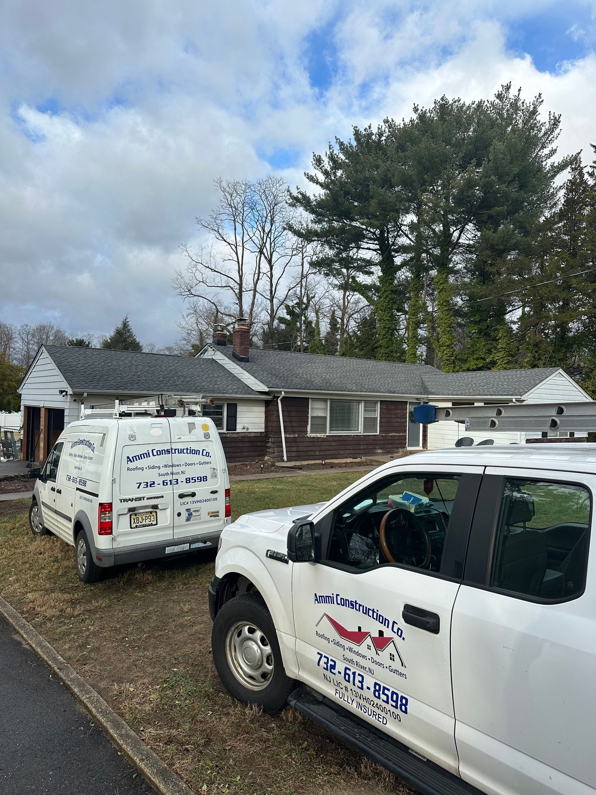 Two white work trucks parked in front of a single-story house with dark siding and a shingled roof under a cloudy sky.