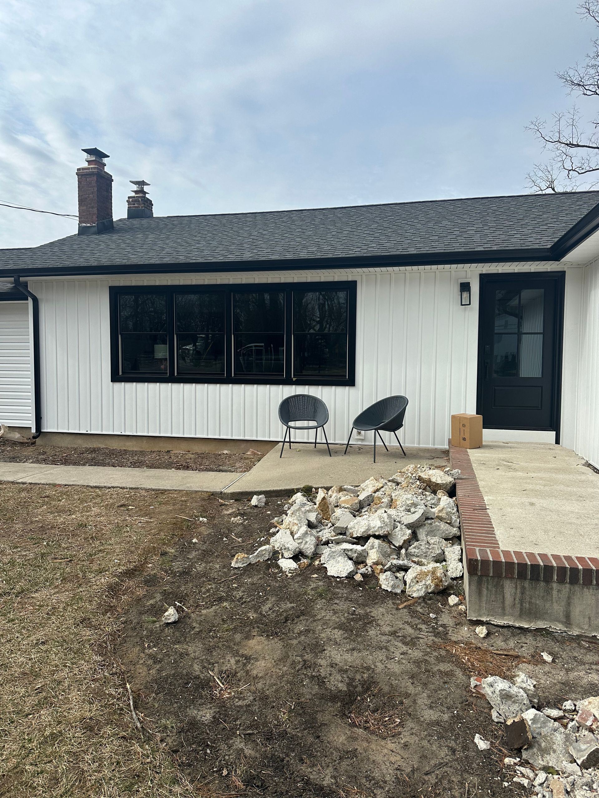 White one-story house with black trim, a large window, a dark front door, and two patio chairs on a concrete walkway.