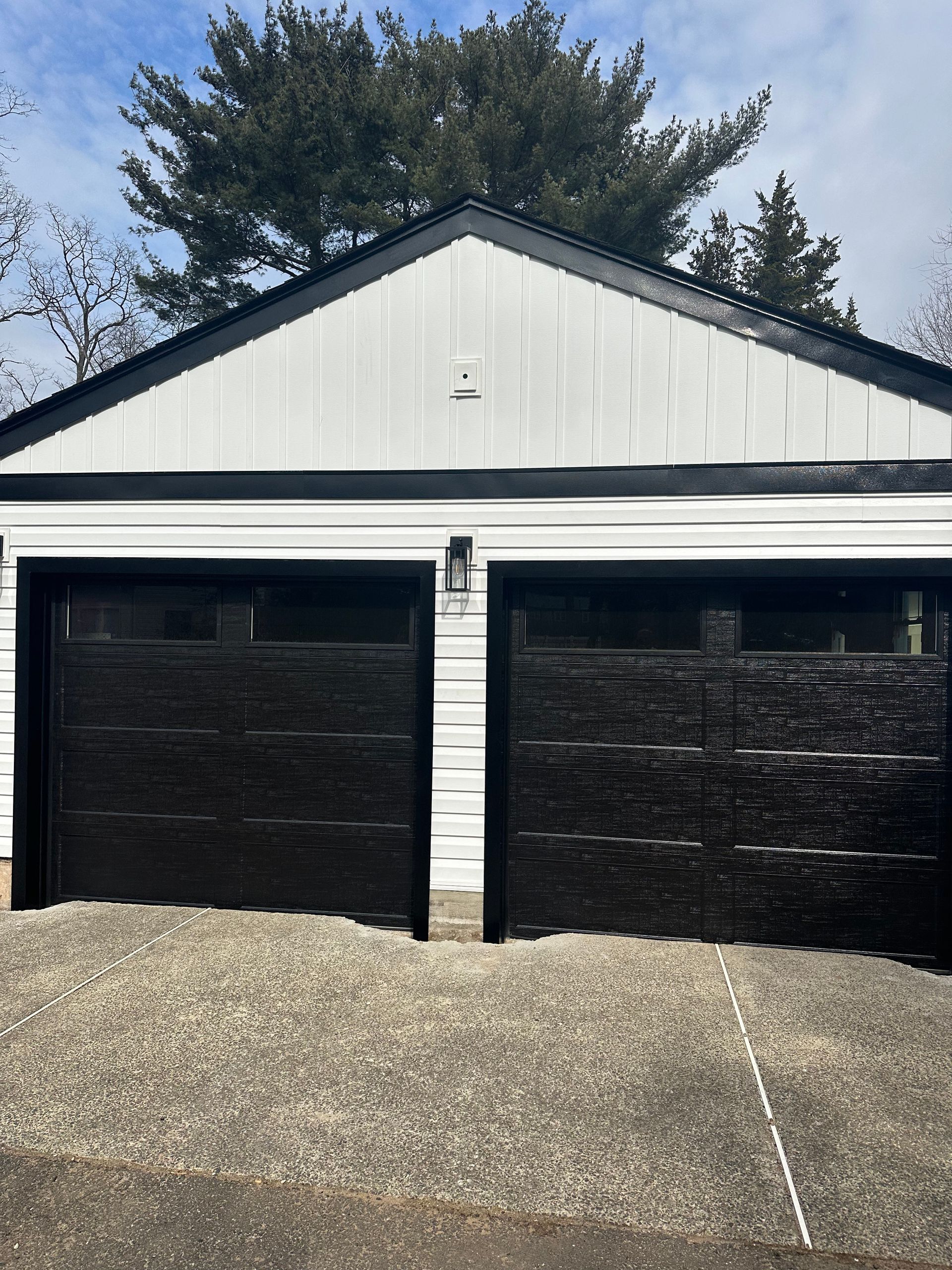 A detached garage with white vertical siding and two black garage doors with rectangular windows, viewed from a driveway.