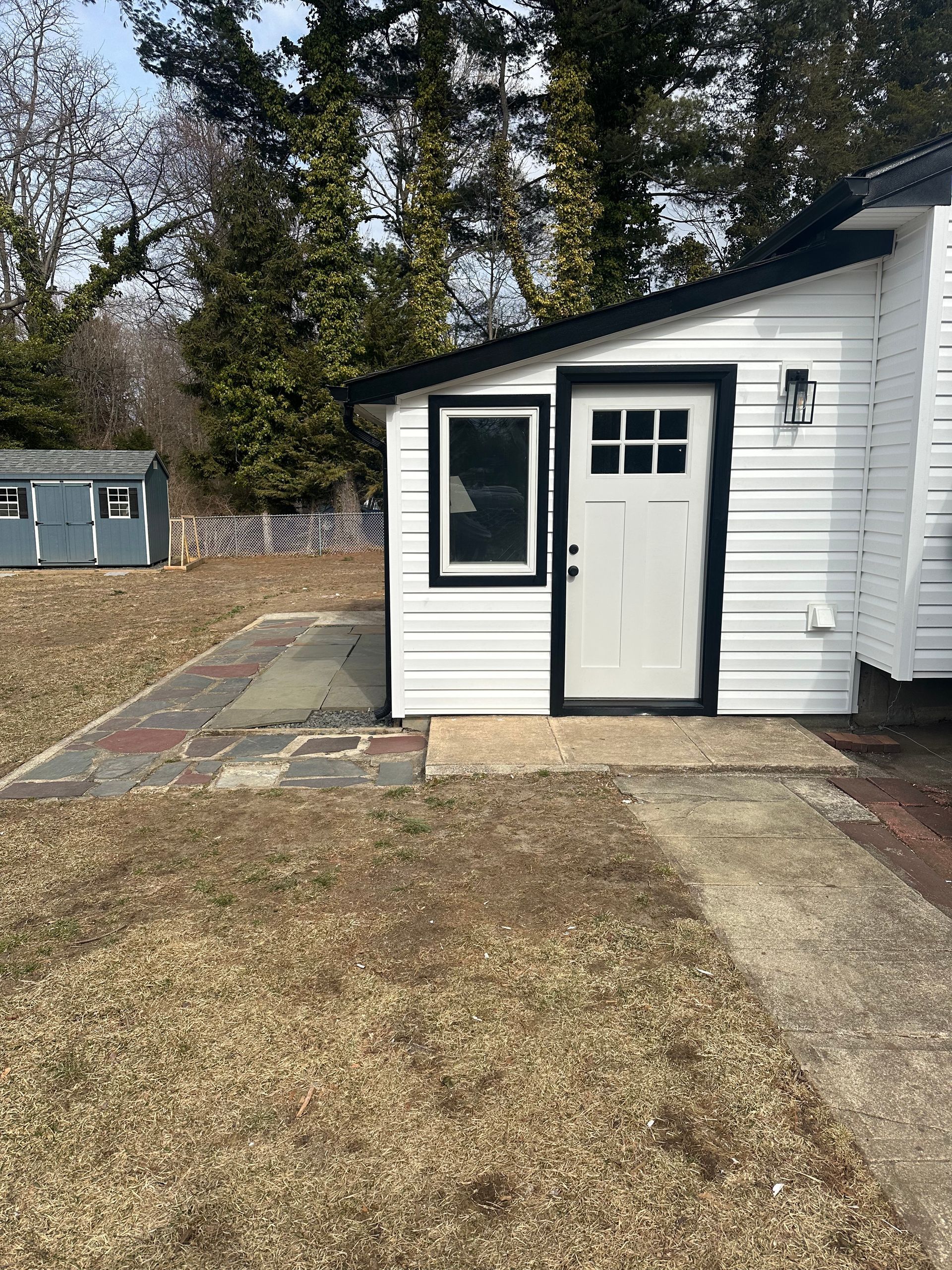 A side view of a white building with a black door and window, featuring a stone patio on a lawn with a shed in the back.