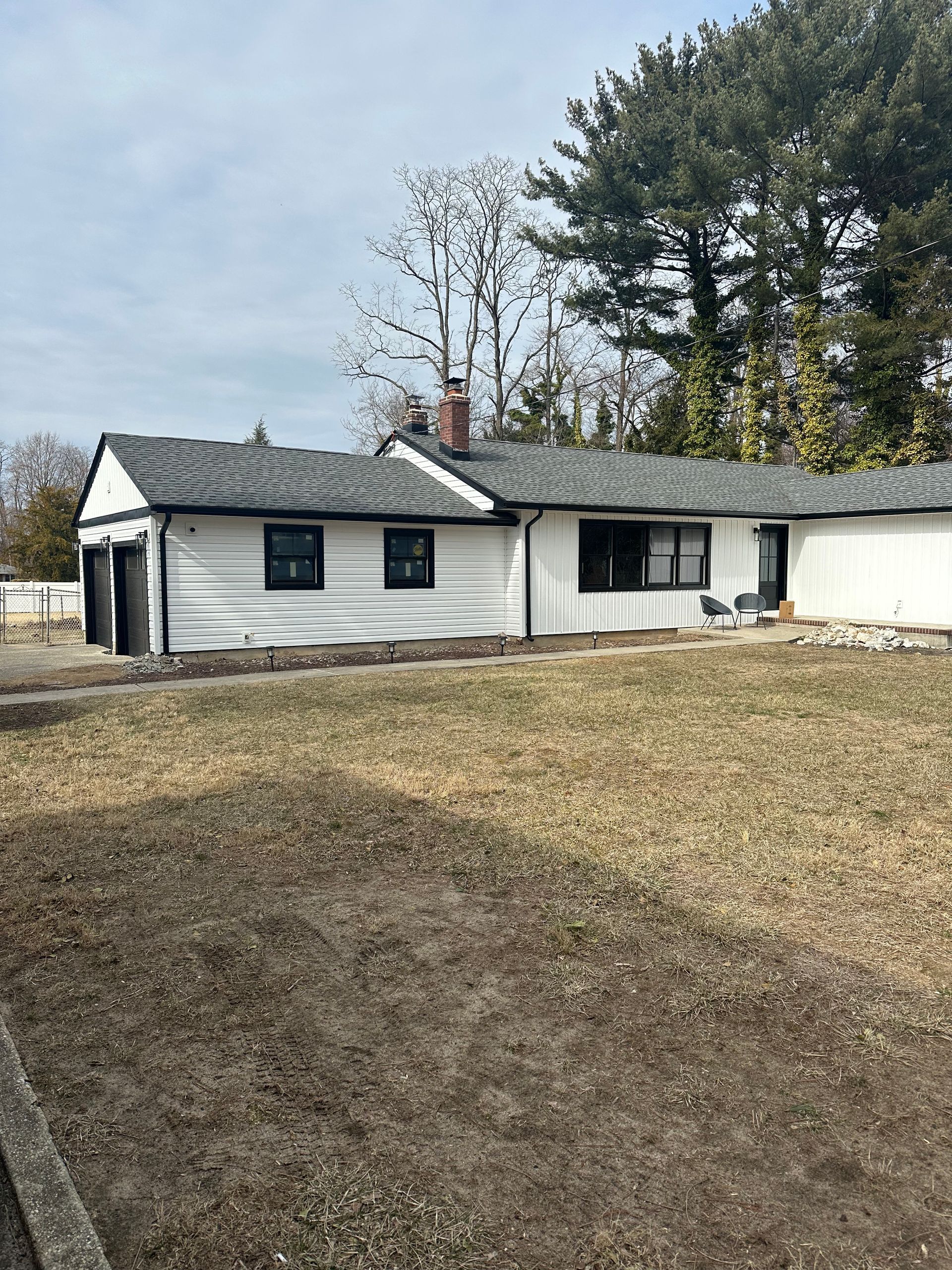 A white, single-story house with a black roof and dark trim stands next to a large yard with sparse, dormant grass.