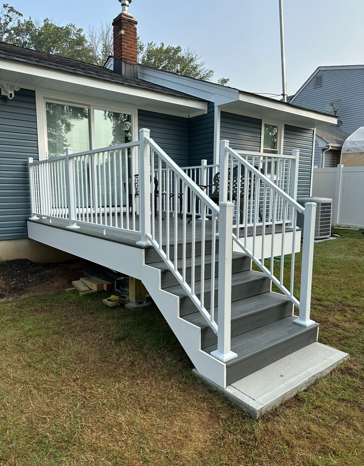 A white vinyl deck and staircase with gray planks attached to the back of a blue-sided house with a brick chimney.