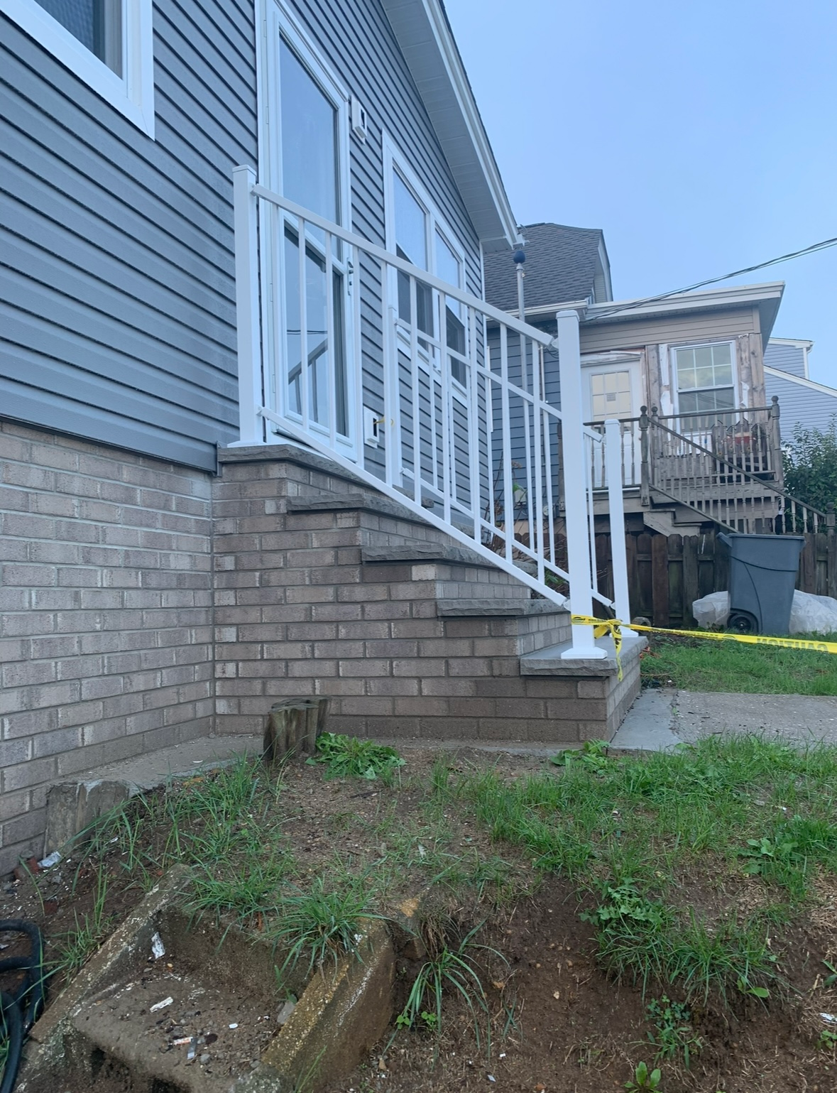 A house entrance with brick stairs and a white metal railing leading to a glass door, with yellow caution tape nearby.
